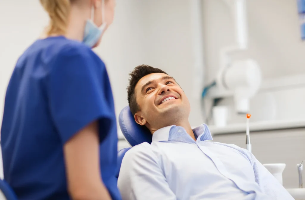 A smiling man in a dental chair talking with a clinician in blue scrubs
