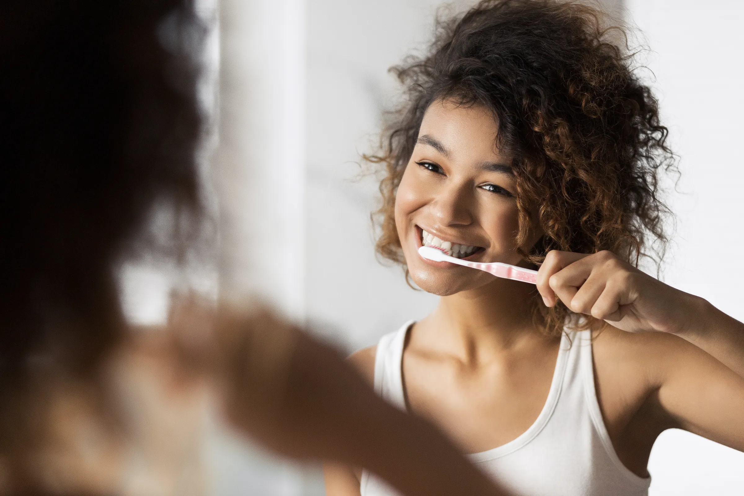 A woman smiling as she brushes her teeth at a bathroom mirror