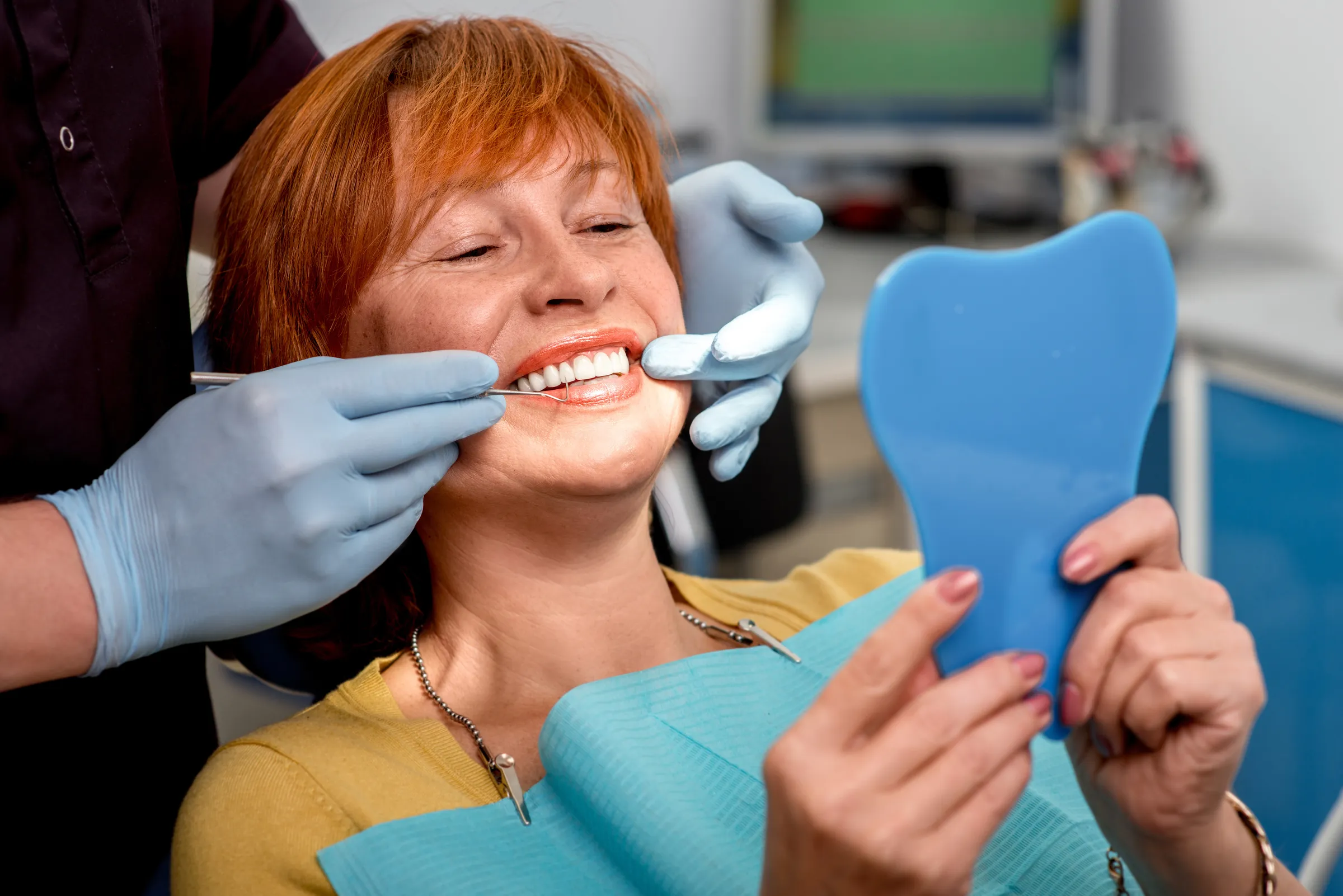 Woman in a dental chair holding a mirror while the dentist checks her new teeth