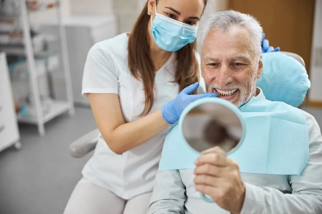 Older man in a dental chair holding a mirror while an assistant looks on