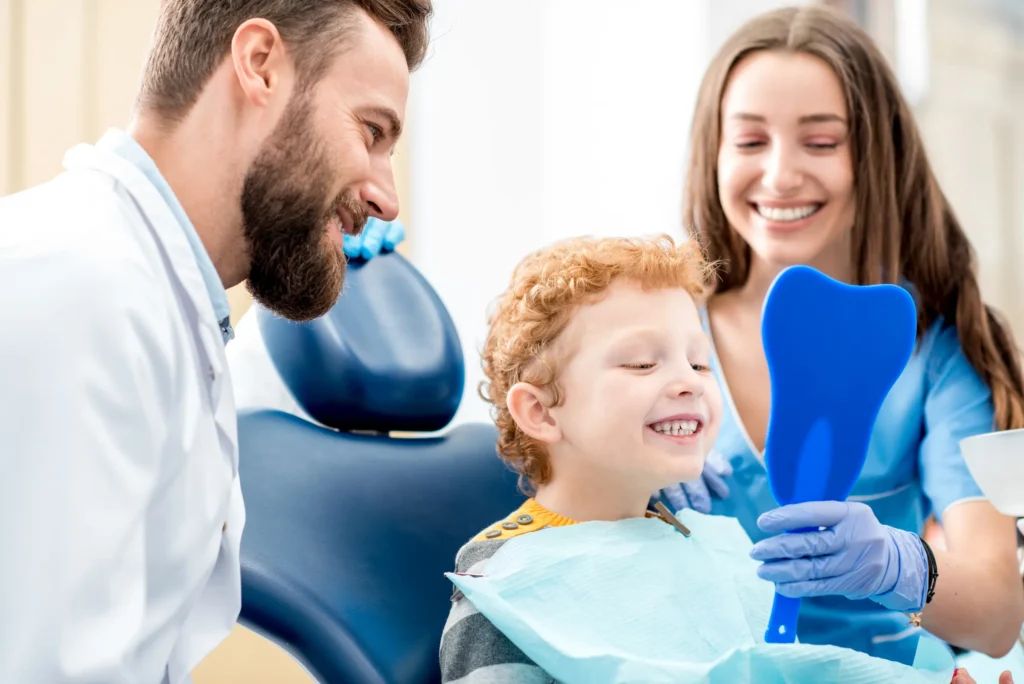 A young boy in a dental chair holding a mirror and smiling, with a dentist beside him