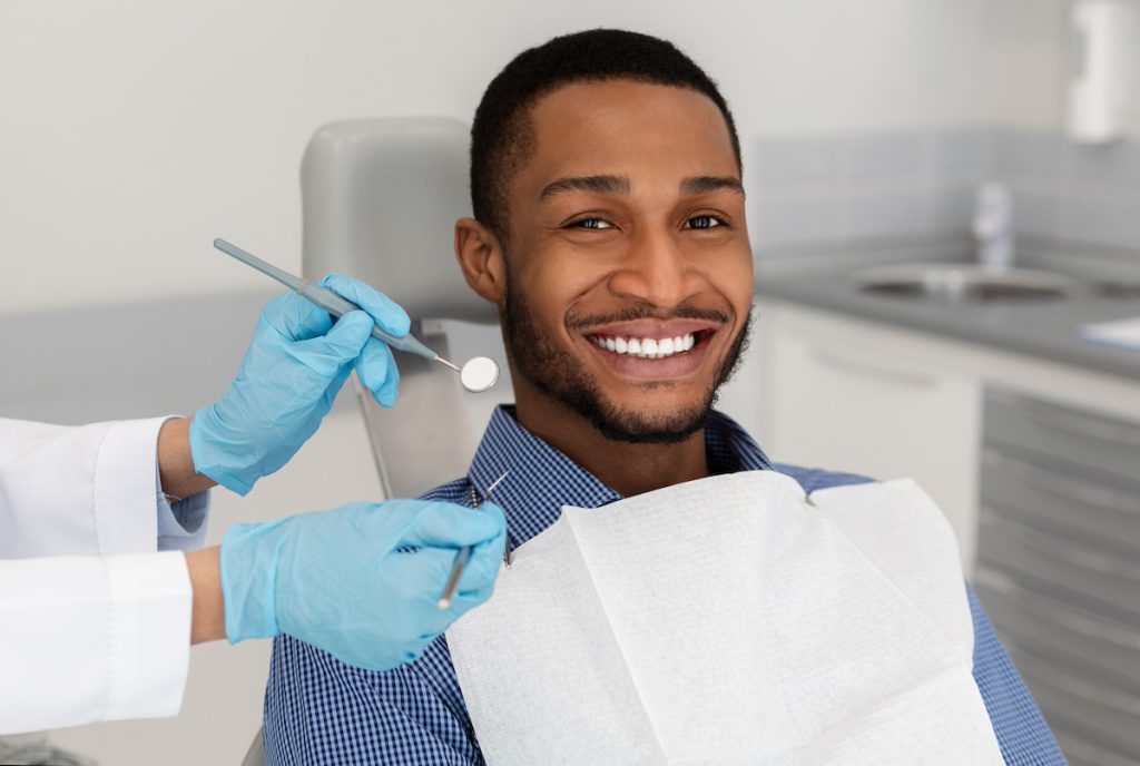 A smiling man in a dental chair while a gloved dentist holds a mirror and probe