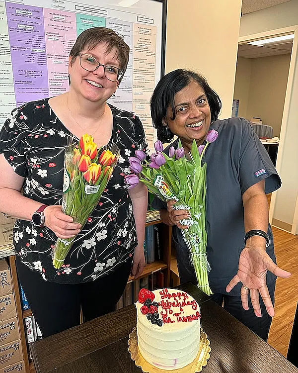 Two team members holding birthday flowers beside a decorated cake