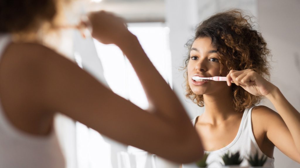 A woman brushing her teeth at a bathroom mirror, seen in the reflection