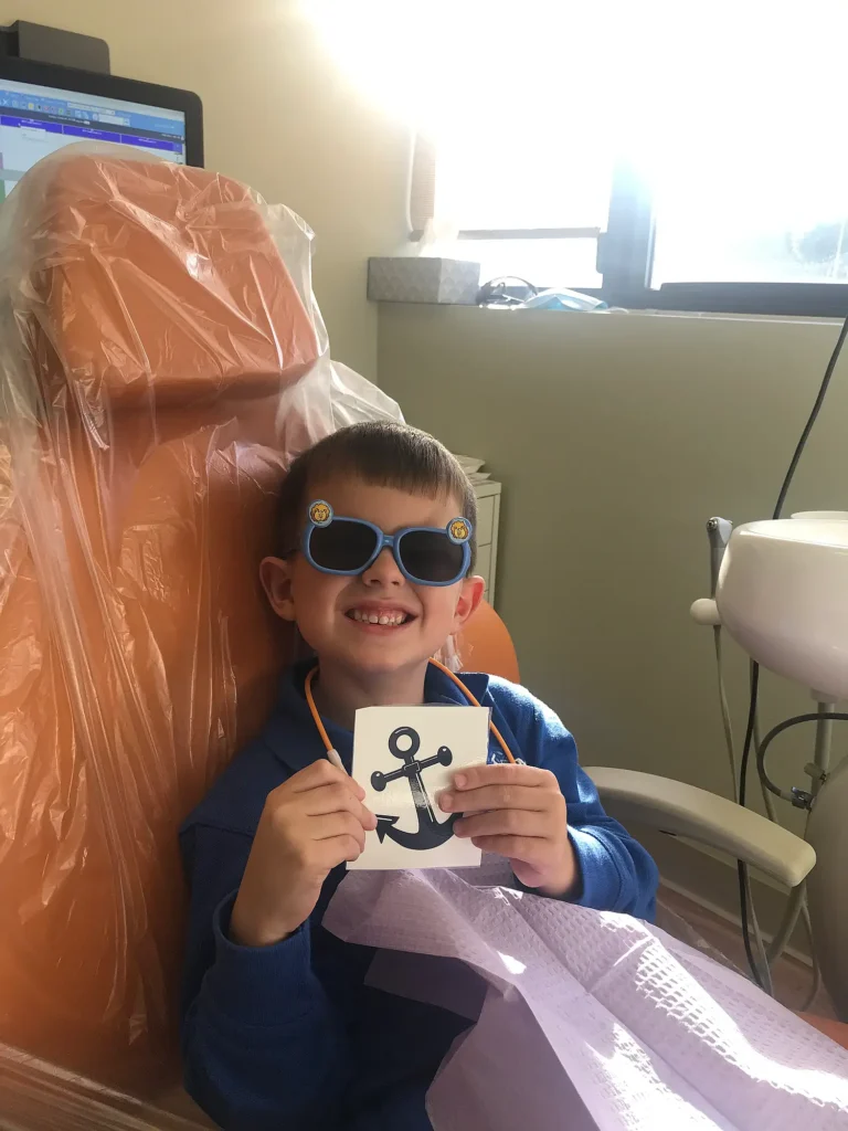 Smiling young patient in sunglasses holding an anchor sticker in the dental chair at MVP Family Dental