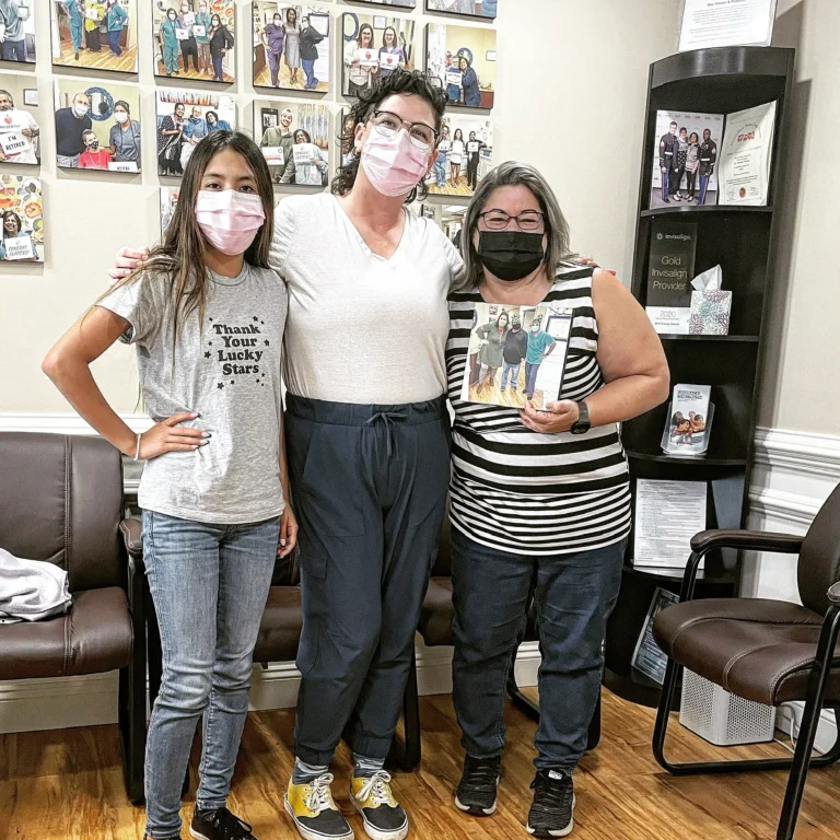 Three patients smiling together in the MVP Family Dental waiting room in front of the patient photo wall