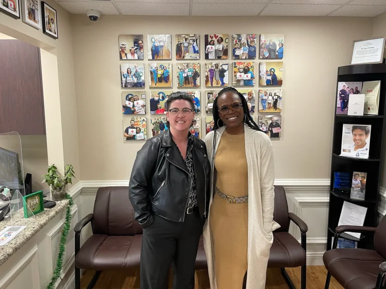 Two smiling patients in front of the patient photo wall at MVP Family Dental