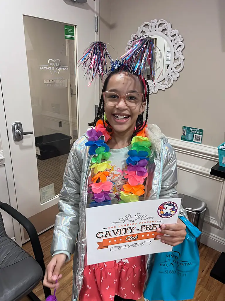 Young patient in a colorful lei and tinsel headband holding a Cavity-Free Club certificate